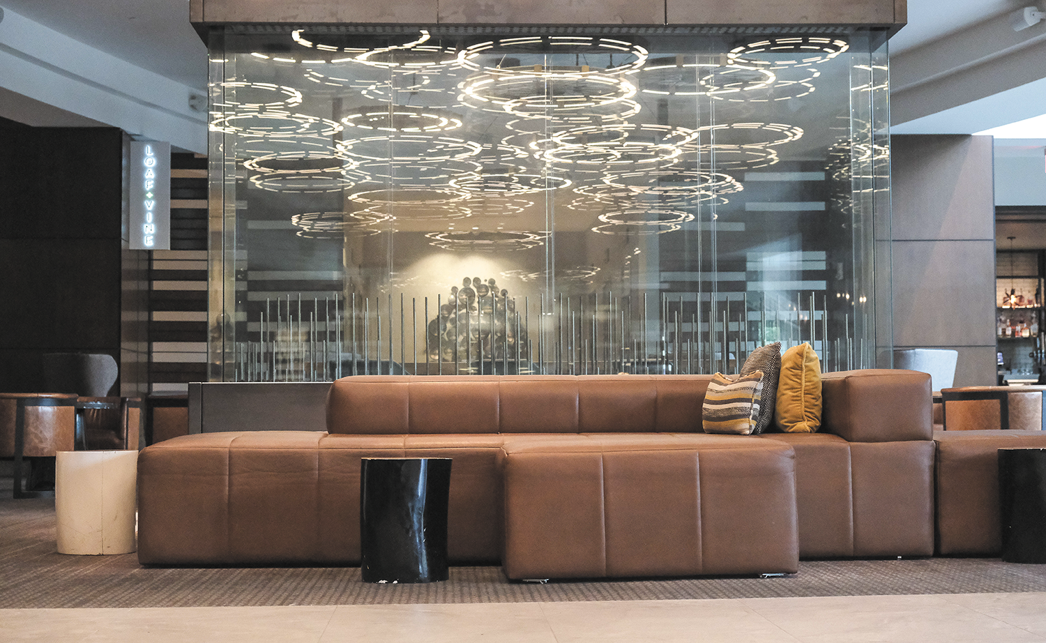 Photograph of an upscale hotel lobby with a light brown leather low back sectional in the foreground. A large panel of glass behind the sofa reflects many overlapping circles of light from a large hanging lighting installation behind the camera.