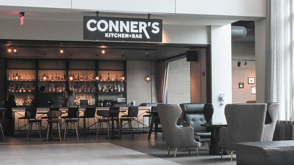 Photograph of a high end hotel bar beneath a hanging sign that reads Conner's Kitchen and Bar. Scattered tables and chairs sit in front of a white subway tiled back bar