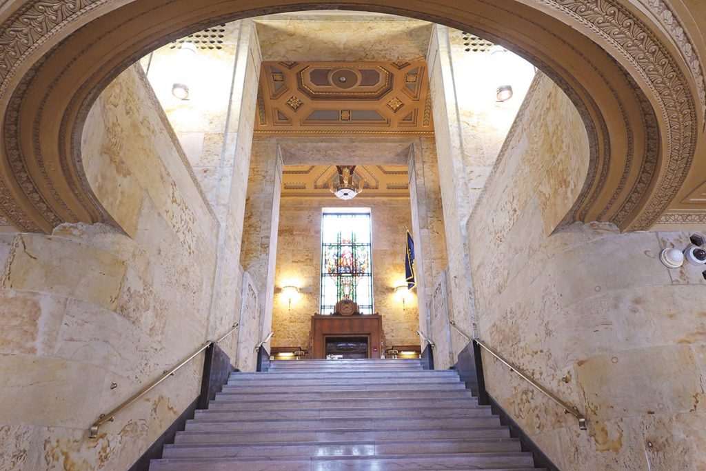 View up a large stairway with a leaded glass window at the top, all of the walls are paneled with stone